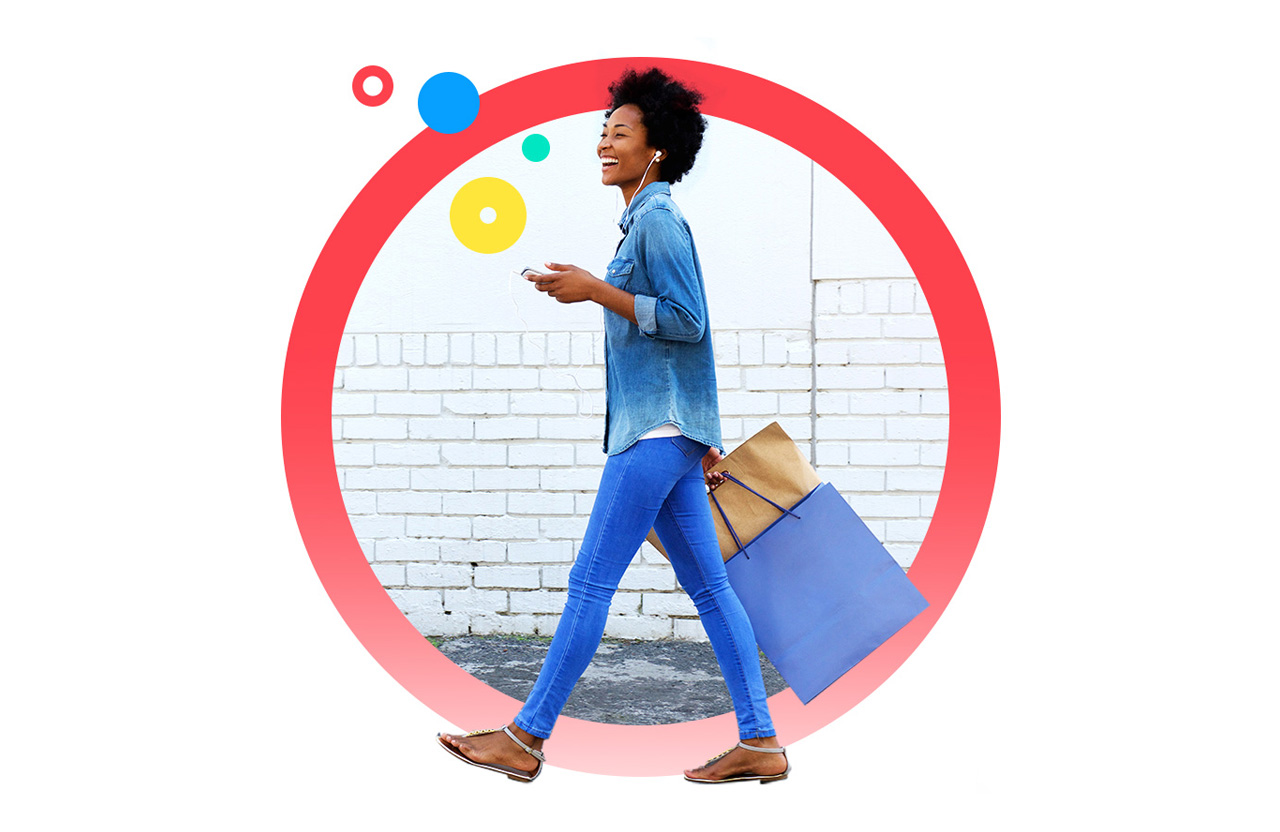 Woman smiling as she walks with shopping bag, phone in hand and wearing earbuds
