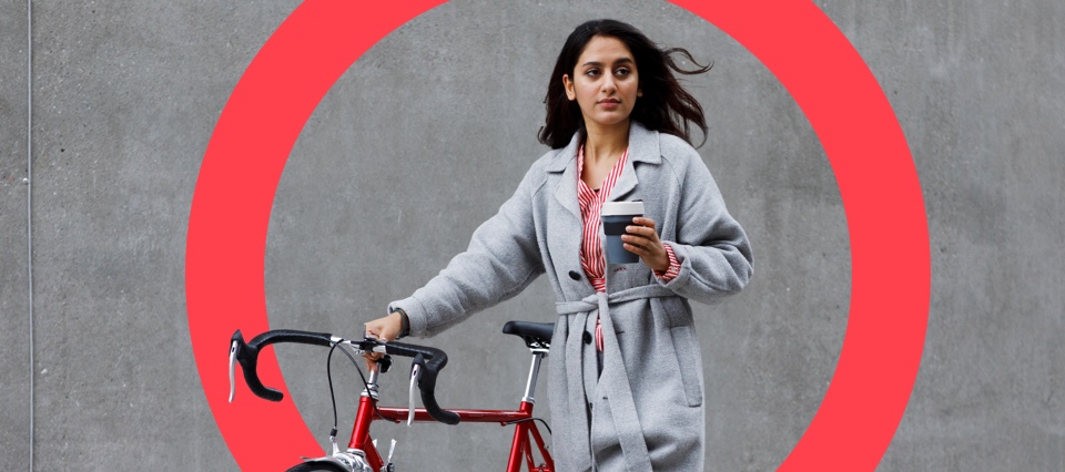 A woman in a grey coat with a bicycle, holding a cup of coffee in front of a large red circle.