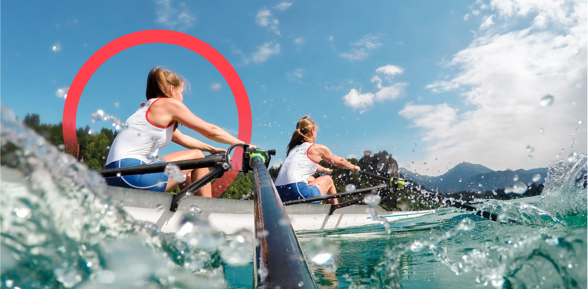 Two female crew members rowing a fast rowing boat in nature with trees and mountains in the background. The camera is positioned on one of the oars with water splashing.
