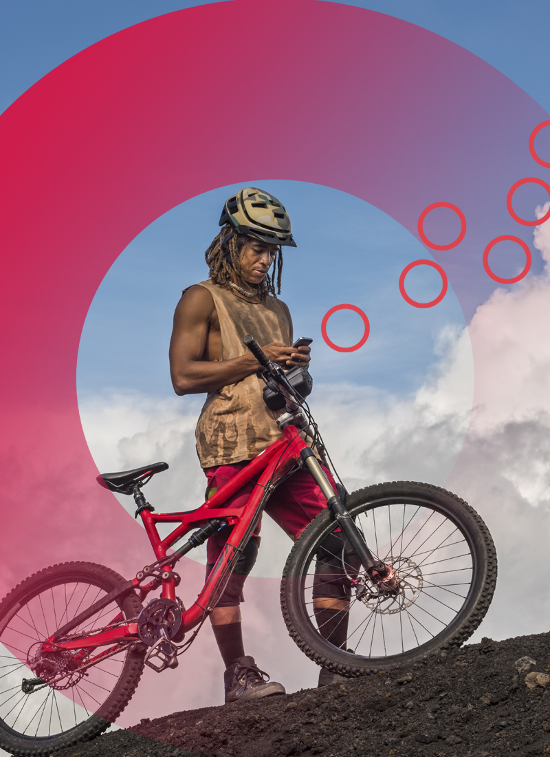 Man wearing athletic equipment and bike helmet standing next to mountain bike on hill looking at phone in hands