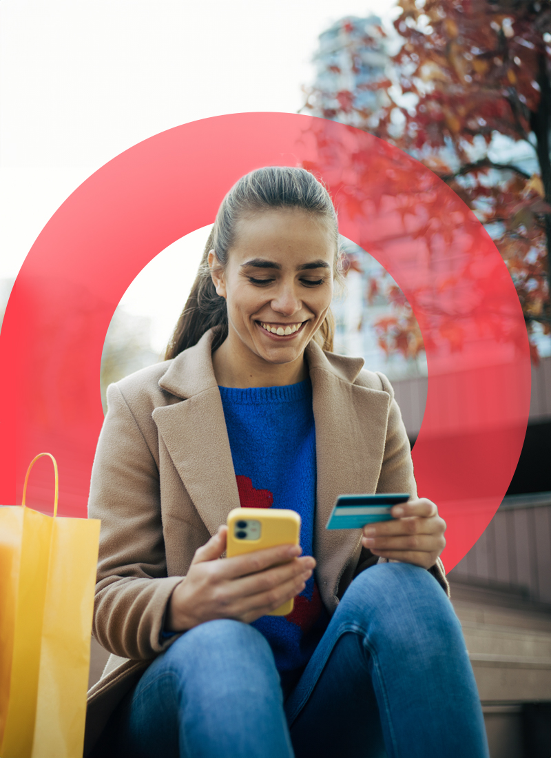 Woman sitting down next to shopping bags and smiling looking at credit card in left hand and phone in right hand.