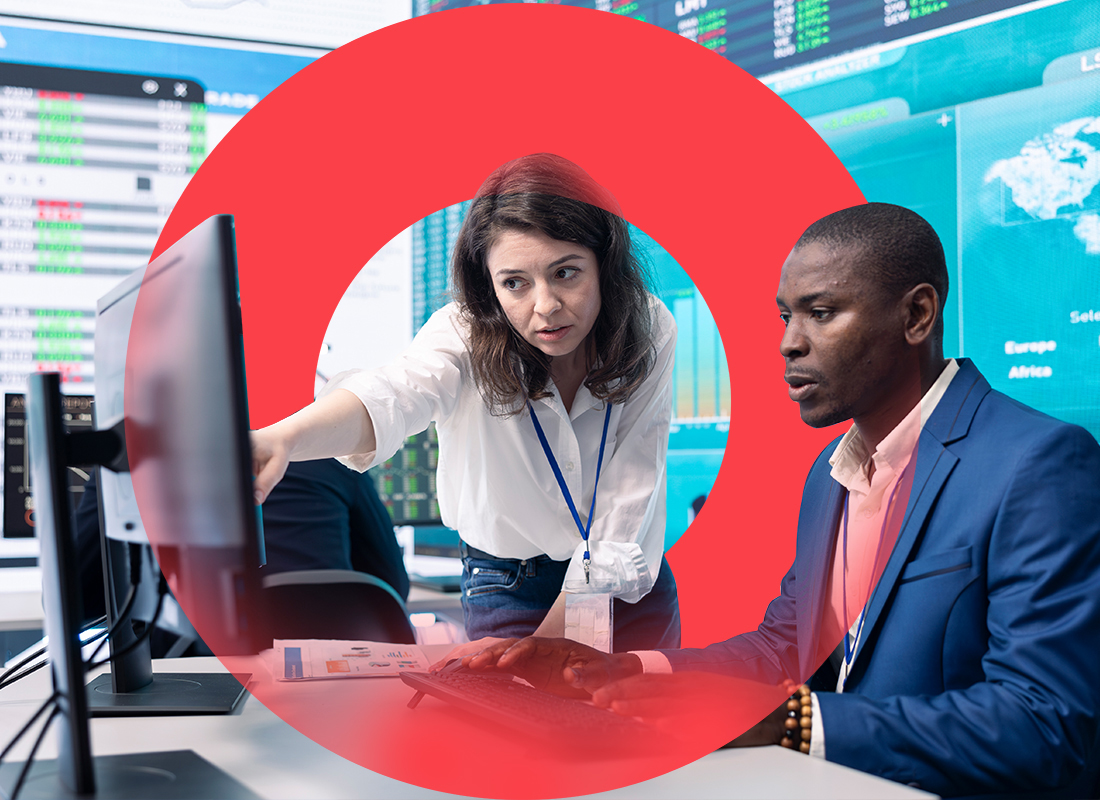 Two business professional collaborating on a computer screen, with a man seated and a woman leaning in and pointing at monitor. 