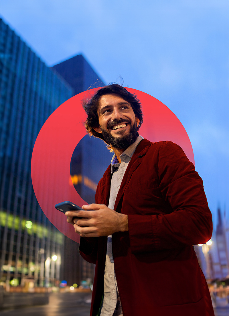 Businessman in a suit standing with a building in the background, smiling, holding a smartphone, and looking upwards