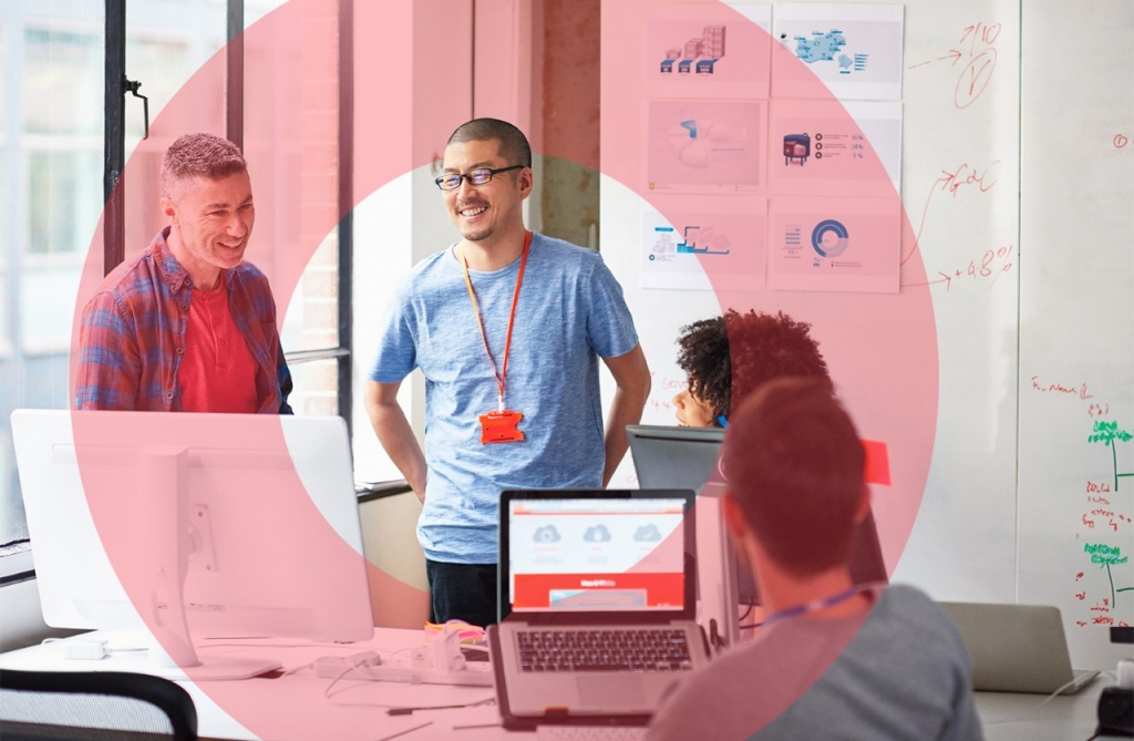 Group of individuals standing and sitting in a work environment with laptops and a computer monitor.