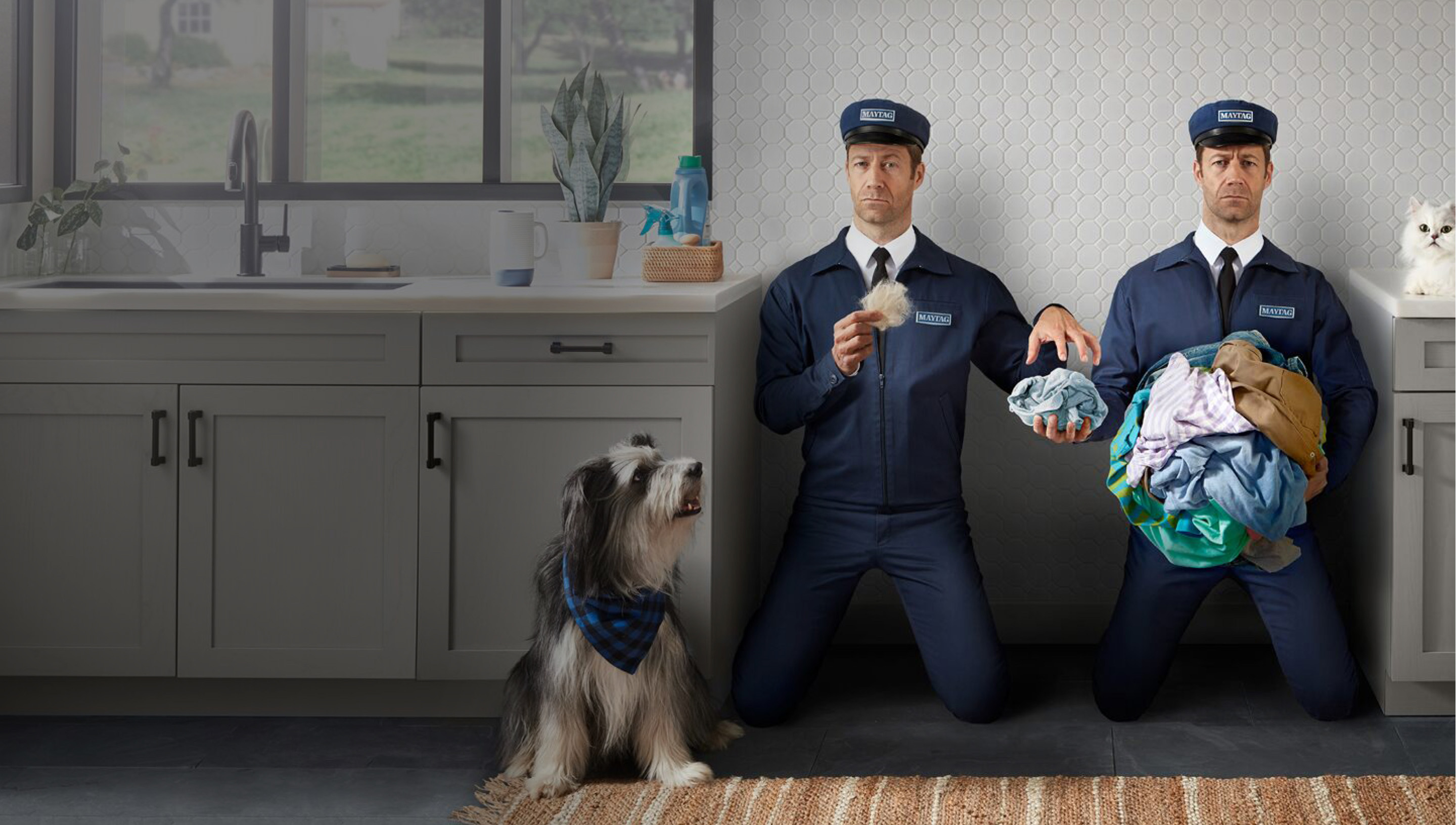 Two male men dressed in Maytag service uniform keeling side by side holding laundry with a shaggy dog looking at them