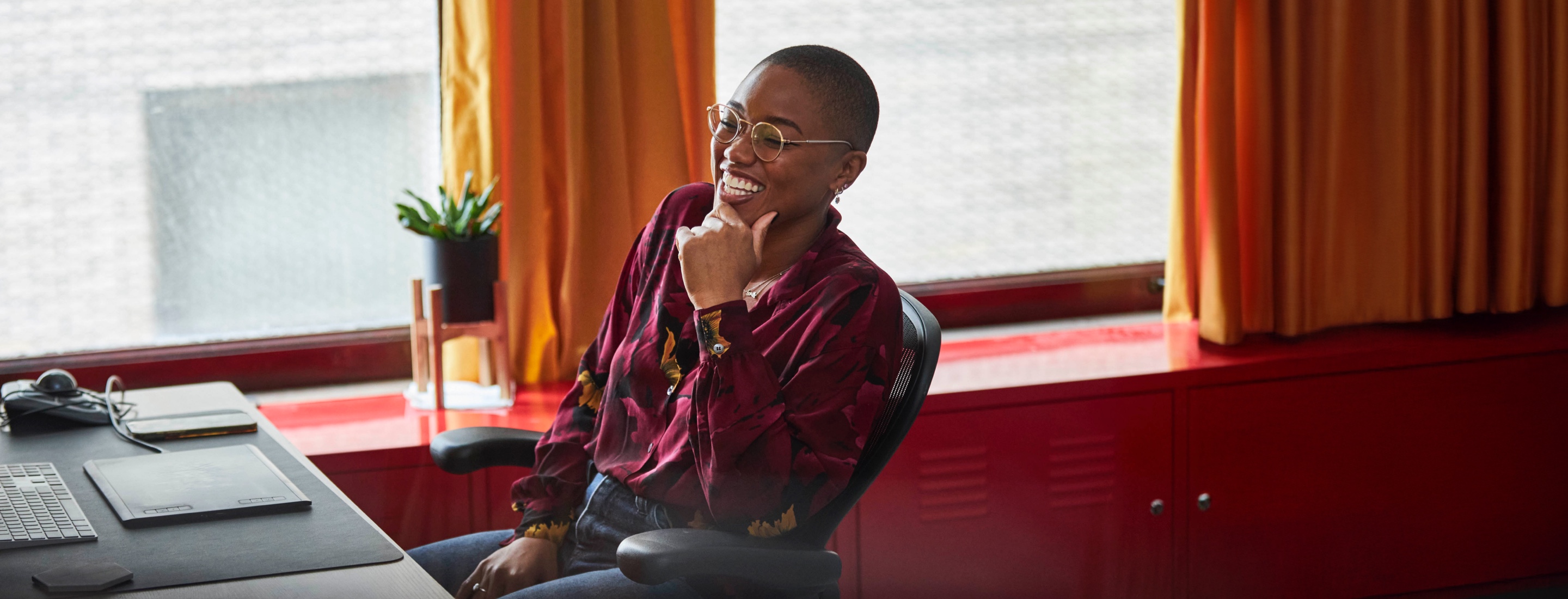 A woman sitting at her work desk places a hand on her chin and smiles.
