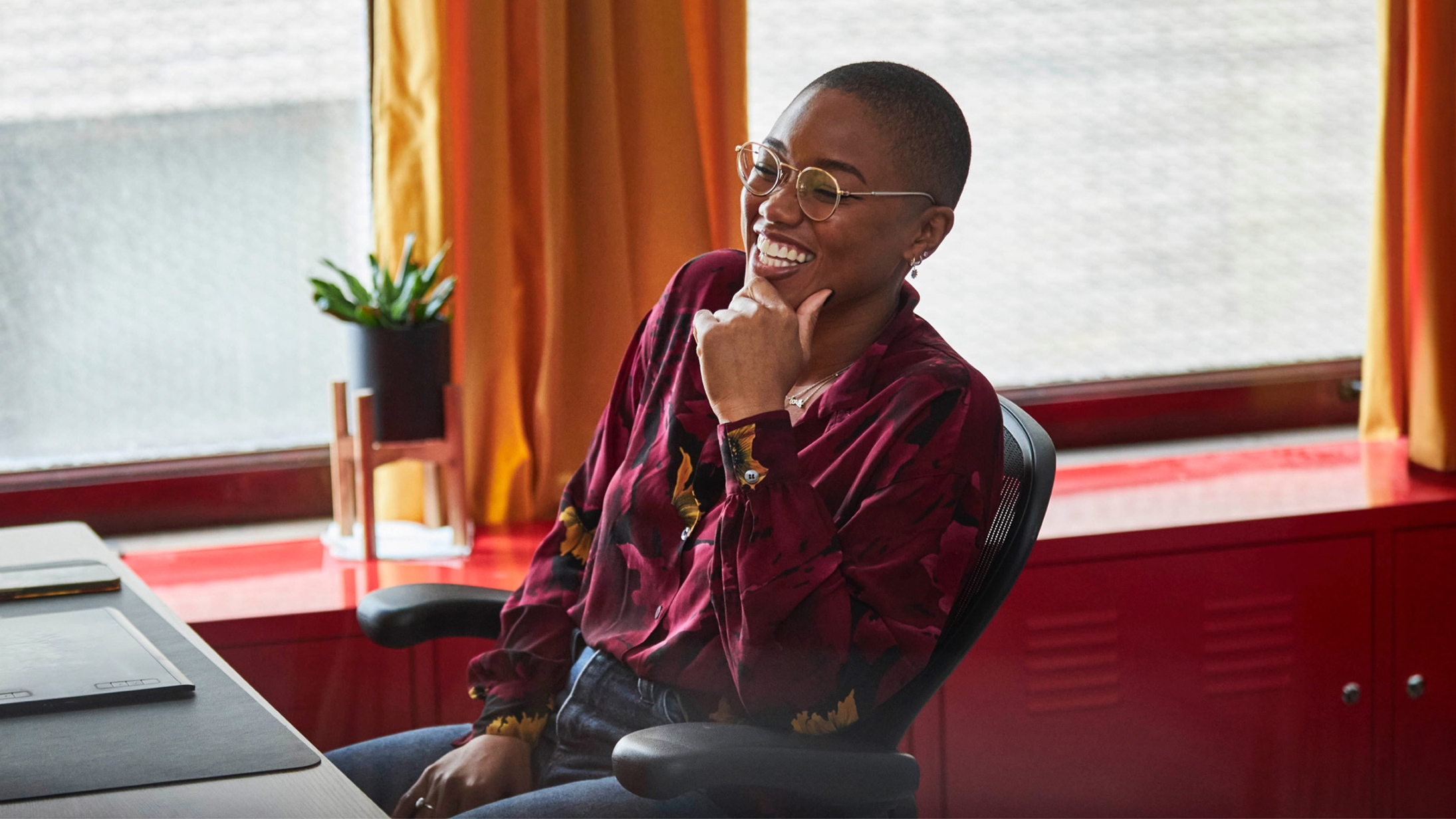 A woman sitting at her work desk places a hand on her chin and smiles. 