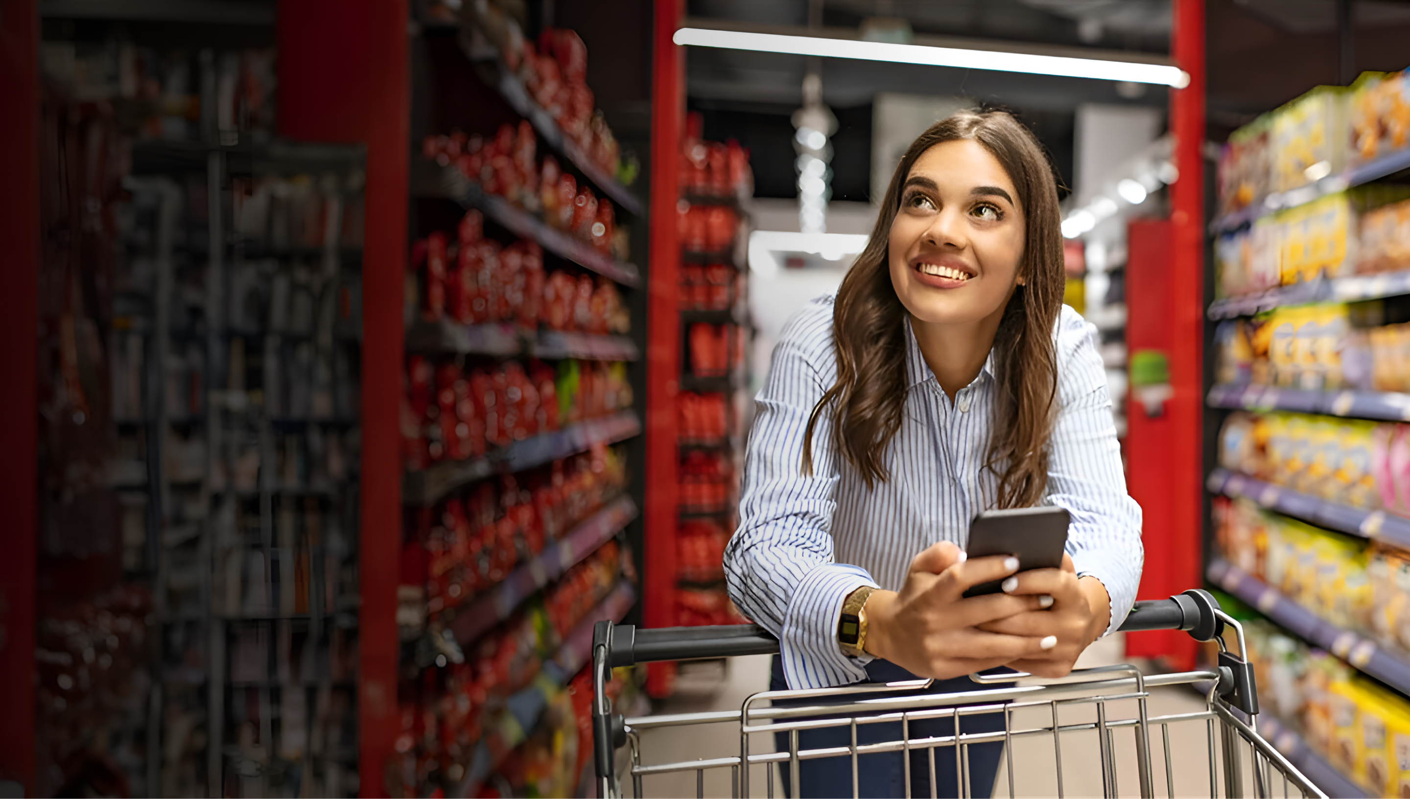 Smiling woman leaning on shopping cart holding phone in grocery aisle