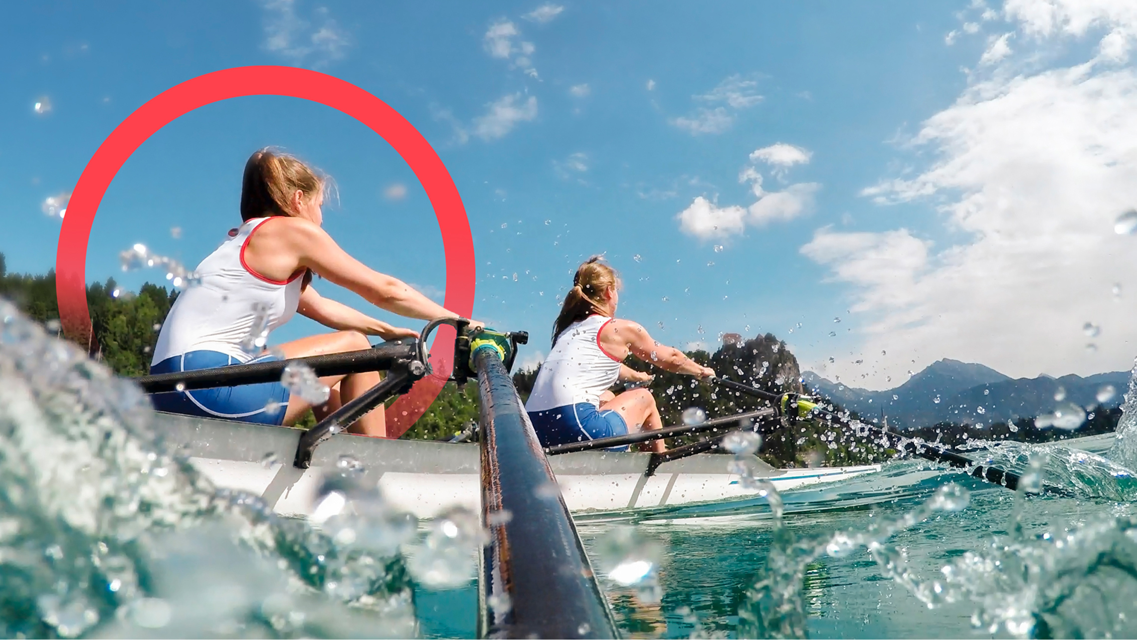 Two female crew members rowing a fast rowing boat in nature with trees and mountains in the background. The camera is positioned on one of the oars with water splashing.