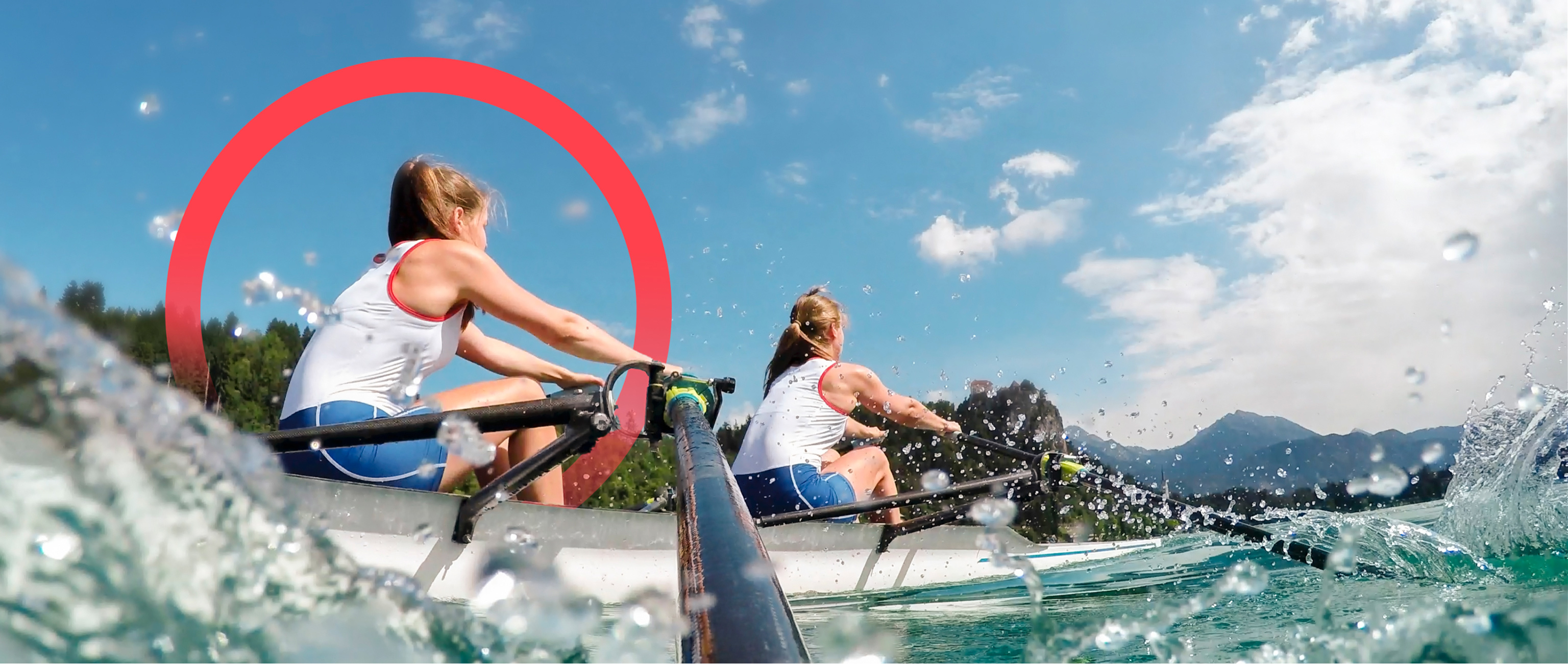 Two female crew members rowing a fast rowing boat in nature with trees and mountains in the background. The camera is positioned on one of the oars with water splashing.
