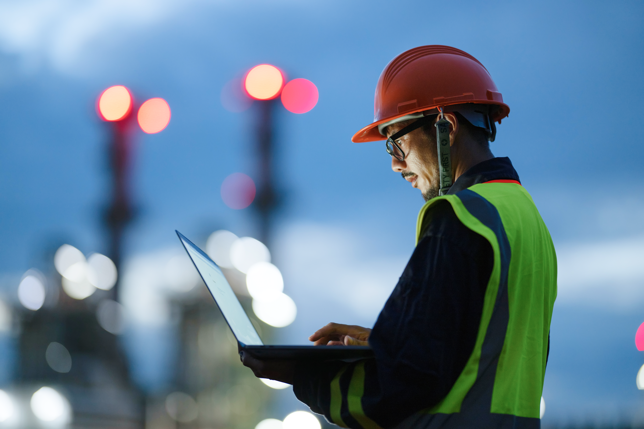 A man wearing a hard hat and safety vest is focused on using a laptop at a construction site.